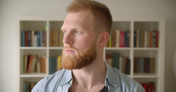 Gros plan portrait de rousse barbu caucasien mâle étudiant regardant caméra dans la bibliothèque — Video