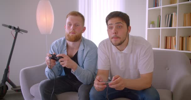 Primer plano retrato de dos hombres caucásicos guapos adultos jugando videojuegos para divertirse sentado en el sofá en el interior — Vídeos de Stock