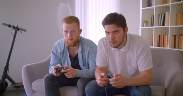 Крупный план портрета двух взрослых красивых белых мужчин, играющих в видеоигры, сидящих на диване в помещении — стоковое видео