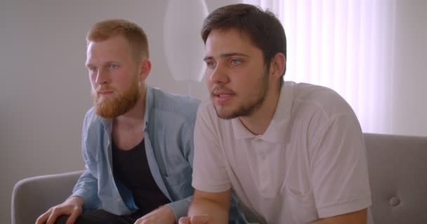 Retrato de cerca de dos hombres caucásicos guapos adultos viendo deportes en la televisión y animando a su equipo de fútbol sentado en el sofá dentro — Vídeos de Stock