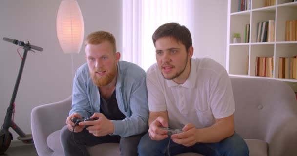 Retrato de cerca de dos hombres caucásicos guapos adultos jugando videojuegos divirtiéndose sentados en el sofá dentro — Vídeos de Stock