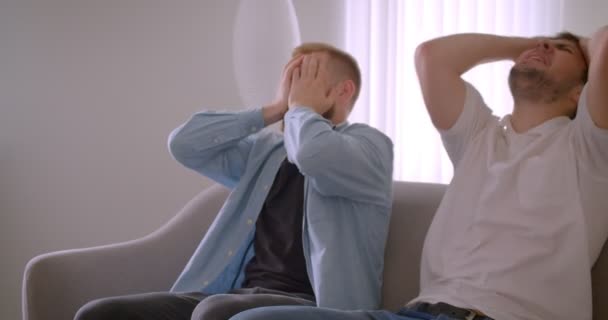 Gros plan portrait de deux adultes beaux hommes caucasiens regarder le sport à la télévision devient triste assis sur le canapé à l'intérieur — Video