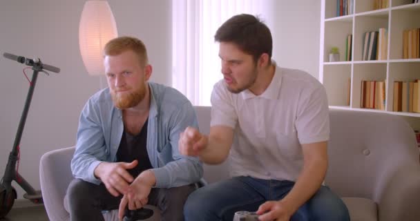 Портрет крупным планом двух взрослых счастливых белых мужчин, играющих в видеоигры, сидящих на диване в помещении — стоковое видео