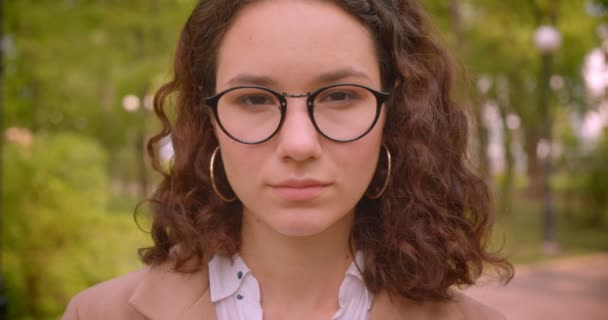 庭に立つカメラを見ながら陽気に微笑むメガネをかけた若い長髪の巻き毛白人女子学生のクローズアップポートレート — ストック動画