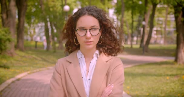 Closeup portré fiatal, hosszú hajú, göndör kaukázusi női hallgató a szemüvegét a karját átkelt mellkasi nézett kamera magabiztosan állva a szabadban a kertben — Stock videók