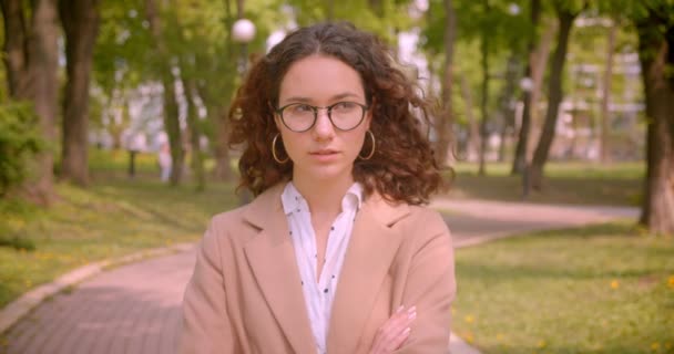 Retrato de cerca de una joven estudiante caucásica rizada de pelo largo en gafas con los brazos cruzados sobre el pecho mirando a la cámara con confianza al aire libre en el jardín — Vídeos de Stock