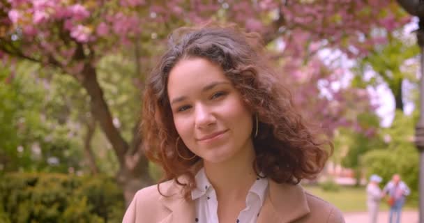 Nahaufnahme Porträt der jungen langhaarigen lockigen Kaukasierin, die glücklich in die Kamera lächelt, die draußen im Park steht — Stockvideo