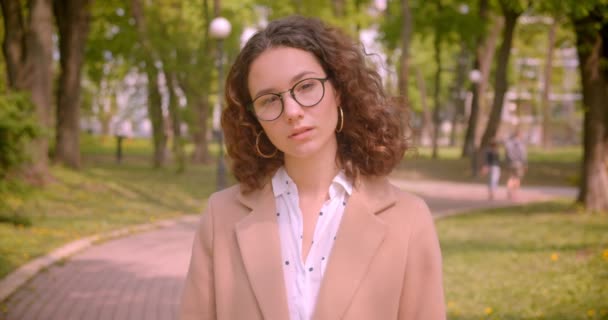 Gros plan portrait de jeune étudiante caucasienne bouclée aux cheveux longs dans des lunettes avec les bras croisés sur la poitrine regardant la caméra debout à l'extérieur dans le jardin — Video