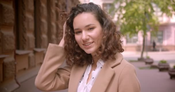Close-up shoot van jonge langharige krullend blanke vrouwelijke student glimlachend vrolijk kijken naar camera staande buitenshuis op straat — Stockvideo