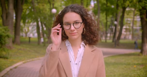 Κοντινό πλάνο του νεαρού μακριά μαλλιά σγουρά καυκάσιος γυναίκα φοιτητής χαμογελάει ευχαρίστως φτιάχνοντας τα γυαλιά της κοιτάζοντας την κάμερα στέκεται σε εξωτερικούς χώρους στο πάρκο — Αρχείο Βίντεο
