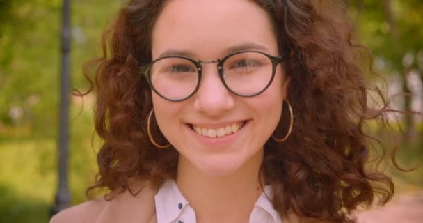 Close-up portret van jonge langharige krullend Kaukasische vrouwelijke student in glazen glimlachend graag naar camera staande buiten in de tuin — Stockvideo