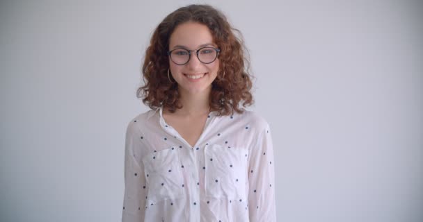 Primer plano de la joven elegante de pelo largo rizado hembra caucásica en gafas sonriendo alegremente mirando a la cámara con el fondo aislado en blanco — Vídeo de stock