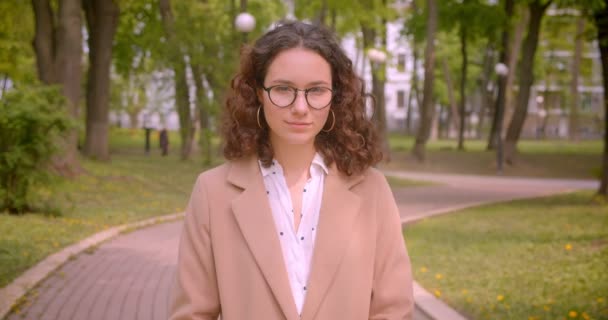 Close-up portret van jonge langharige krullend Kaukasische vrouwelijke student in glazen kijken naar camera staande buiten in de tuin — Stockvideo