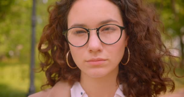 Close-up portret van jonge langharige krullend Kaukasische vrouwelijke student in glazen kijken naar camera staande buiten in de tuin — Stockvideo