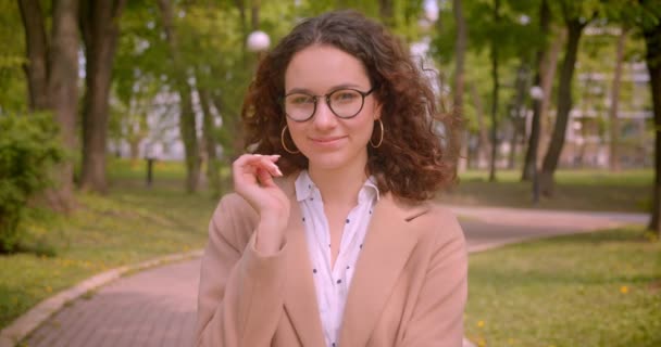 Retrato de cerca de una joven estudiante caucásica rizada de pelo largo sonriendo felizmente arreglando sus gafas mirando a la cámara al aire libre en el parque — Vídeos de Stock