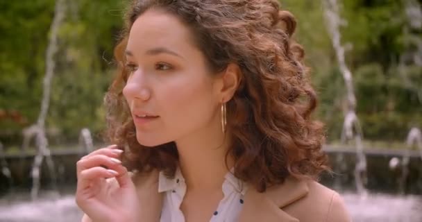 Closeup portrait of young beautiful pretty long haired curly caucasian female looking at camera outdoors in the garden with fountains on the background — Stock Video