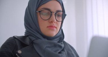 Ofis kapalı bir koltukta oturan kamera bakarak dizüstü bilgisayar kullanarak hijab ve gözlük genç çekici Müslüman iş kadını Closeup portre