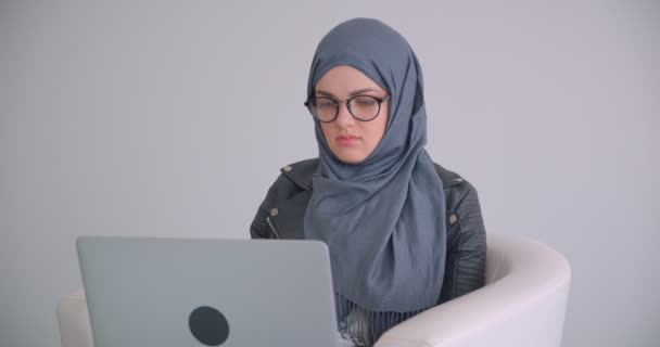 オフィスの屋内のアームチェアに座っているラップトップを使用してヒジャーブとメガネで若い魅力的なイスラム教徒のビジネスウーマンのクローズアップ肖像画 — ストック動画