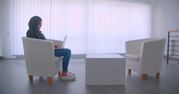 Closeup side view portrait of young attractive muslim businesswoman in hijab and glasses working on the laptop sitting in an armchair indoors in the office — Stock Video