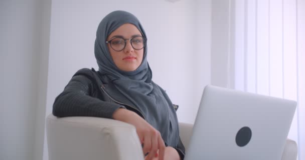 Retrato de close-up de jovem atraente mulher de negócios muçulmana em hijab e óculos com laptop olhando para a câmera sentada em uma poltrona dentro de casa no escritório — Vídeo de Stock