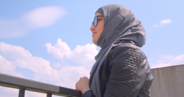 Κοντινό γύρισμα της νεαρής όμορφη μουσουλμανική θηλυκό σε χιτζάμπ και γυαλιά κοιτάζοντας την αστική θέα της πόλης και γυρίζοντας στην κάμερα χαμογελαστά χαρούμενα — Αρχείο Βίντεο