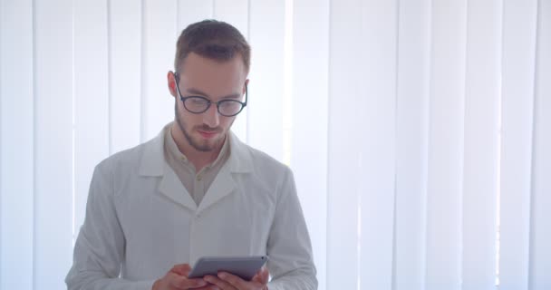 特写镜头中年轻的英俊的白种男医生看着相机微笑着愉快地拿着平板电脑在白色的公寓 — 图库视频影像