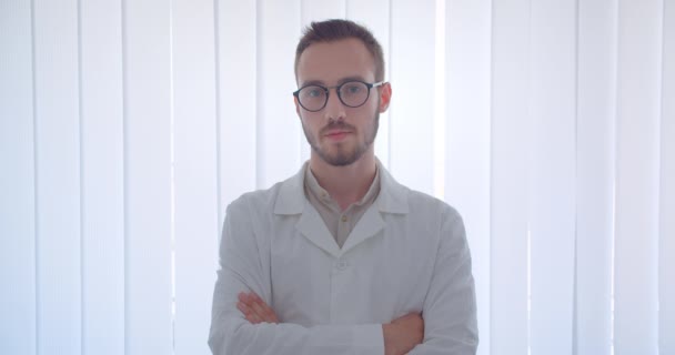 Close-up portret van jonge knappe Kaukasische mannelijke arts kijken naar camera glimlachend gelukkig met zijn armen gekruist over de borst in het witte appartement — Stockvideo