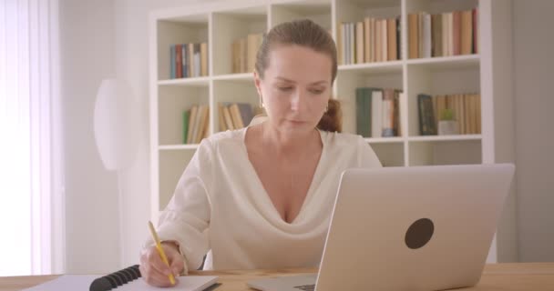 Крупный план портрета пожилой белой брюнетки-предпринимательницы, использующей ноутбук и делающей заметки в офисе в помещении — стоковое видео