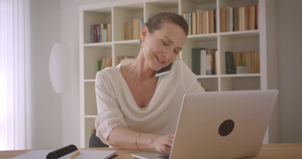 Närbild porträtt av äldre kaukasiska brunett affärskvinna med hjälp av den bärbara datorn och med ett telefonsamtal på kontoret inomhus — Stockvideo