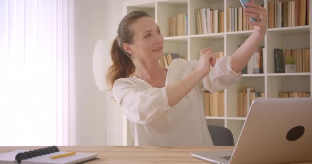 Портрет крупным планом пожилой белой брюнетки-предпринимательницы, делающей селфи по телефону и позирующей перед ноутбуком в офисе в помещении — стоковое видео