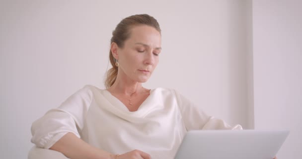 Κοντινό πλάνο των ηλικιωμένων Καυκάσιος μελαχρινή επιχειρηματίας χρησιμοποιώντας το φορητό υπολογιστή κοιτάζοντας την κάμερα κάθεται στην πολυθρόνα σε ένα λευκό διαμέρισμα σε εσωτερικούς χώρους — Αρχείο Βίντεο