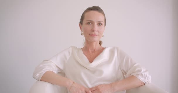 Крупним планом портрет літньої білої брюнетки, яка дивиться на камеру, сидячи в кріслі в білій квартирі в приміщенні — стокове відео