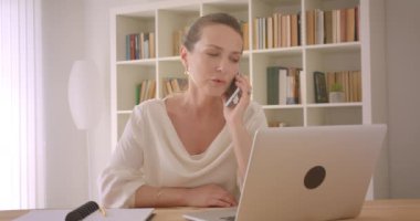 Dizüstü bilgisayar kullanarak ve kapalı ofiste telefonda bir konuşma yaşlı kafkas esmer iş kadını Closeup portre