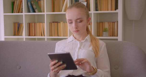 Retrato de close-up de jovem estudante loira muito caucasiana usando o tablet olhando para a câmera sentada no sofá dentro de casa no apartamento — Vídeo de Stock