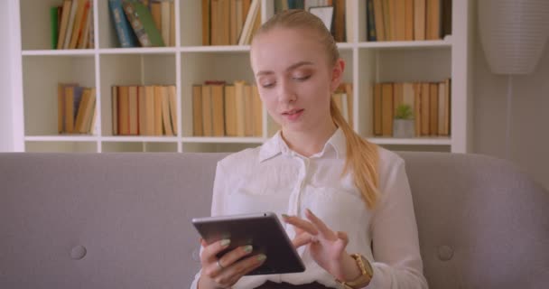 Retrato de close-up de jovem estudante loira muito caucasiana usando o tablet olhando para a câmera sorrindo feliz sentado no sofá dentro de casa no apartamento — Vídeo de Stock