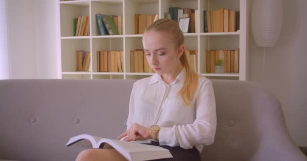 Närbild porträtt av unga ganska kaukasiska blonda kvinnliga studenten läsa en bok och titta på kameran sitter på soffan inomhus i lägenheten — Stockvideo