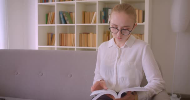 Närbild porträtt av unga ganska kaukasiska blonda kvinnliga student i glasögon läsa en bok och titta på kameran sitter på soffan inomhus i lägenheten — Stockvideo