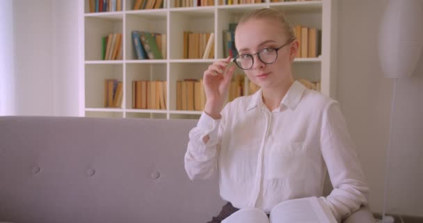 Крупним планом портрет молодої досить кавказької блондинки студентки в окулярах читаючи книгу і дивлячись на камеру посміхаючись весело сидячи на дивані в приміщенні в квартирі — стокове відео