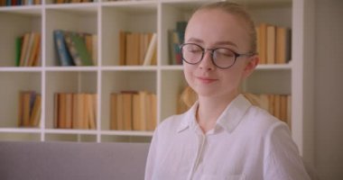 Genç güzel beyaz sarışın kız öğrencinin gözlüklü yakın çekim portresi, dairenin içinde kanepede otururken kameraya gülümseyerek bakıyor