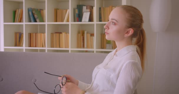 Крупним планом портрет молодої досить кавказької блондинки студентки в окулярах дивиться на камеру посміхаючись весело сидячи на дивані в приміщенні в квартирі — стокове відео