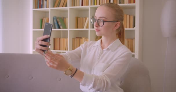 Närbild porträtt av unga ganska kaukasiska blonda kvinnliga student i glasögon tar selfies på telefonen leende glatt sitter på soffan inomhus i lägenheten — Stockvideo