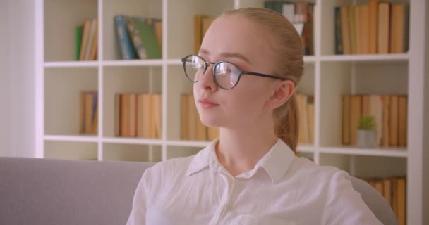 Closeup portrait of young pretty caucasian blonde female student in glasses looking at camera sitting on the sofa indoors in the apartment — Stock Video