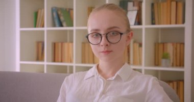 Genç güzel beyaz sarışın kız öğrencinin gözlüklü yakın çekim portresi dairenin iç mekanlarında kanepede otururken kameraya gülümseyerek bakıyor