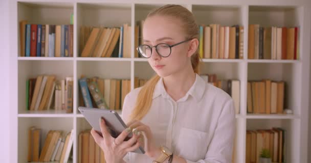 Крупним планом портрет молодої досить кавказької блондинки студентки в окулярах за допомогою планшета і показуючи зелений екран на камеру, що стоїть в приміщенні бібліотеки — стокове відео