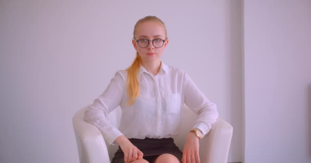 Close-up portret van jonge mooie Kaukasische blonde zakenvrouw in glazen kijken naar camera zittend in de fauteuil binnenshuis in het witte appartement — Stockvideo