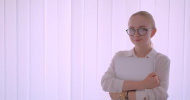 Gözlüklü genç, beyaz sarışın kız öğrencinin, beyaz dairede kapalı alanda duran kameraya neşeyle bakan bir dizüstü bilgisayar tutan yakın çekim portresi