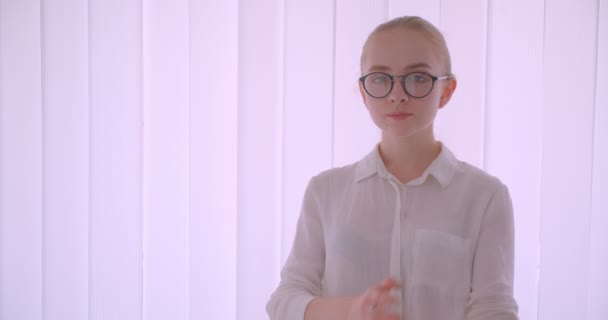 Gros plan portrait de jeune jolie étudiante blonde caucasienne en lunettes regardant la caméra debout à l'intérieur dans l'appartement blanc — Video