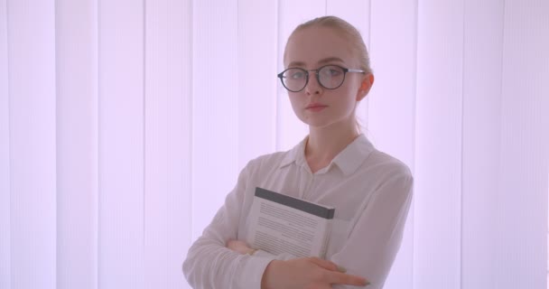 Κοντινό πλάνο του νεαρού όμορφη Καυκάσιος ξανθιά γυναίκα φοιτητής σε γυαλιά κρατώντας ένα βιβλίο κοιτάζοντας την κάμερα στέκεται σε εσωτερικούς χώρους στο λευκό διαμέρισμα — Αρχείο Βίντεο
