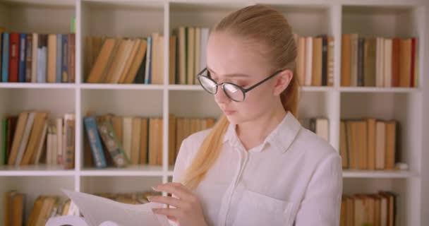 Retrato de cerca de una joven guapa rubia caucásica estudiante de gafas leyendo un libro de pie en la biblioteca con estanterías en el fondo — Vídeo de stock