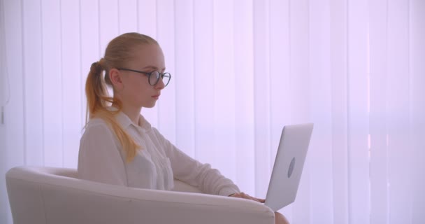 Nahaufnahme Seitenansicht Porträt einer jungen hübschen kaukasischen blonden Geschäftsfrau mit Brille, die den Laptop benutzt und in die Kamera schaut, die im Sessel in der weißen Wohnung sitzt — Stockvideo